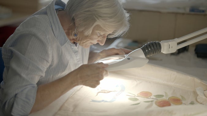 A woman is bent over some white fabric looking through a magnifying glass with a light attached